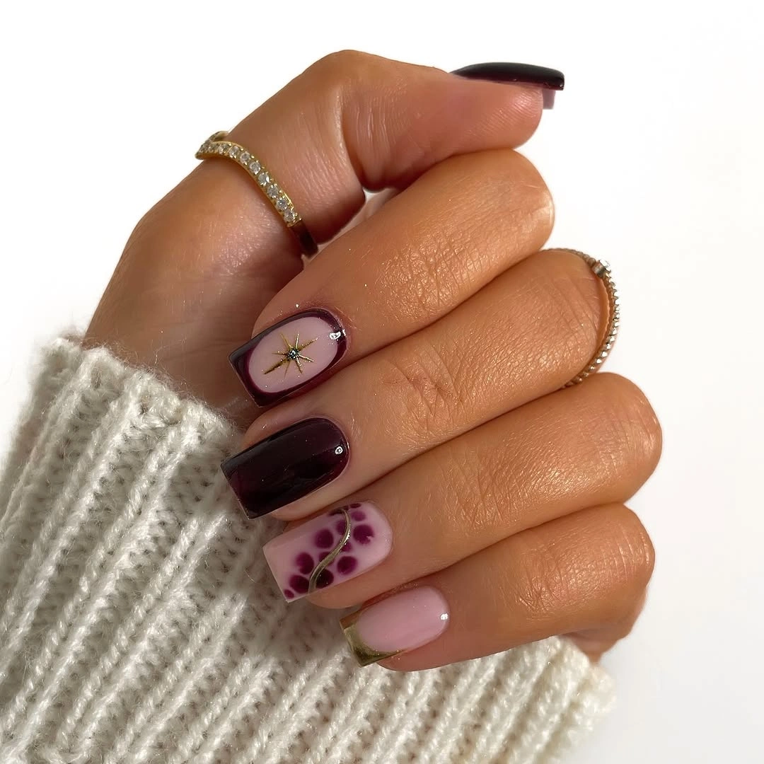 short square manicure featuring burgundy polish, blush accents, gold starburst, and berry dots
