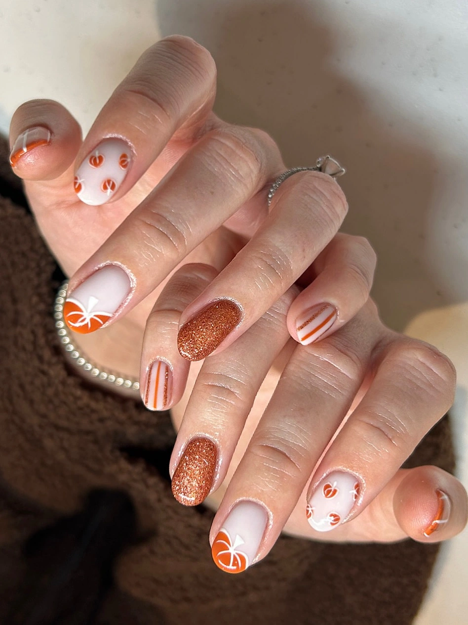 short rounded manicure featuring pumpkin orange motifs, white base accents, stripes, and copper glitter nails