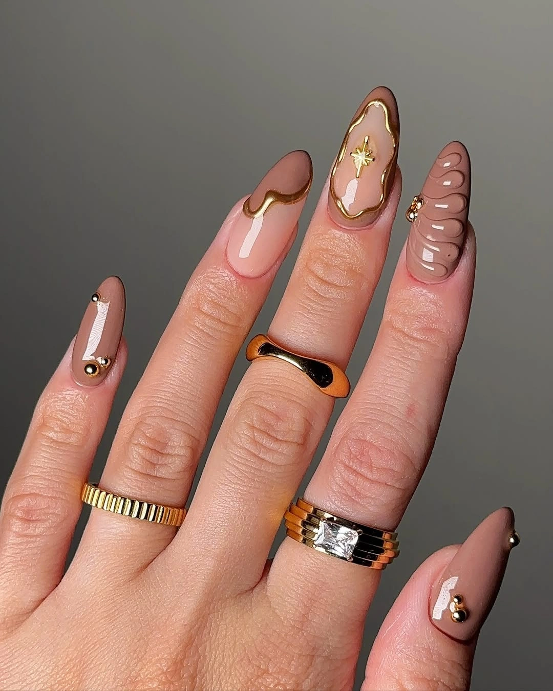 almond manicure featuring taupe nude bases, raised-looking gold curves, star accent, and small beads
