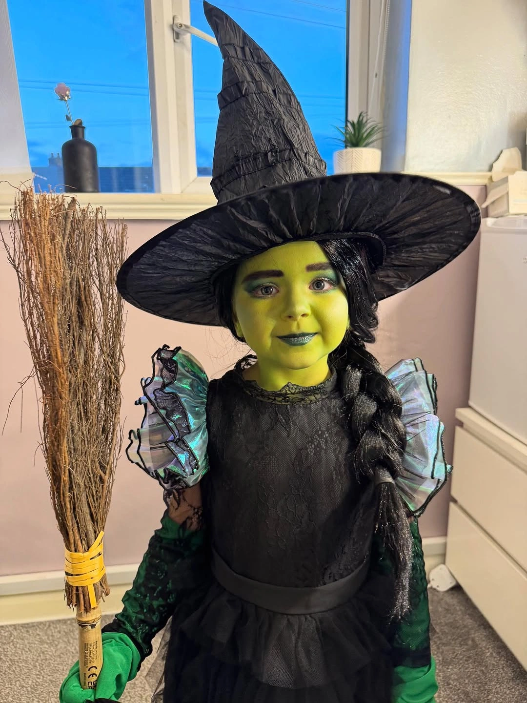 Kids green-faced witch costume with oversized black pointed hat dark dress and green sleeve detail