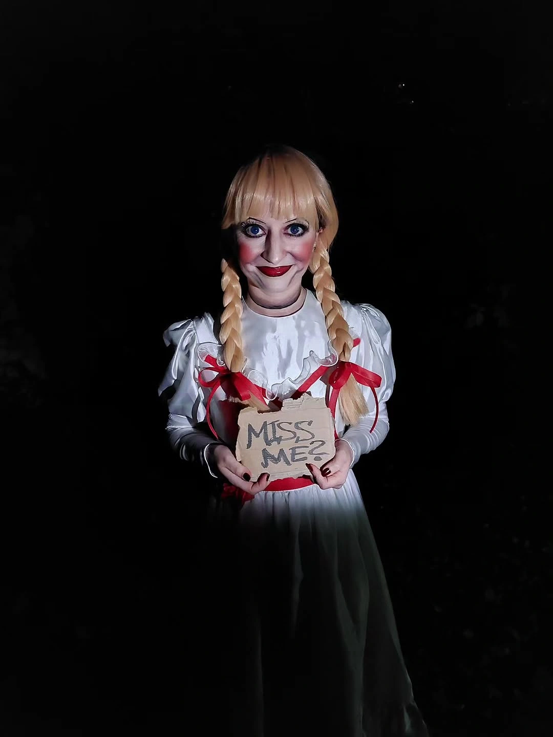 White ruffled horror doll costume with red ribbon accents and handheld sign