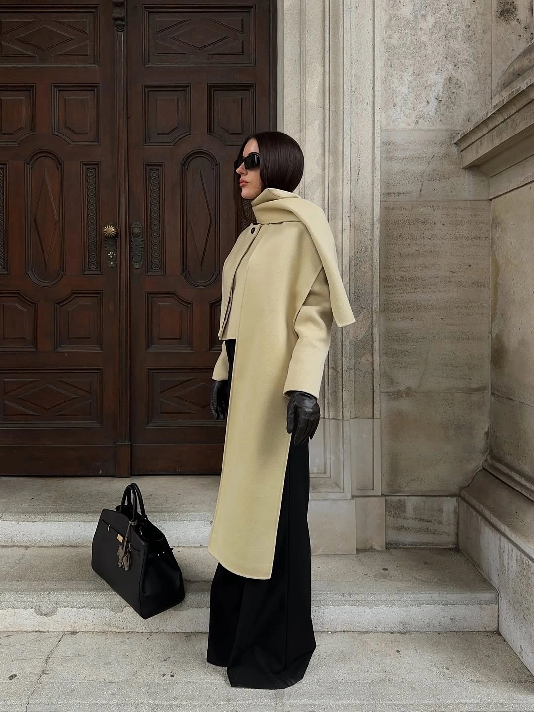 Pale long coat with black wide trousers, gloves, and structured bag