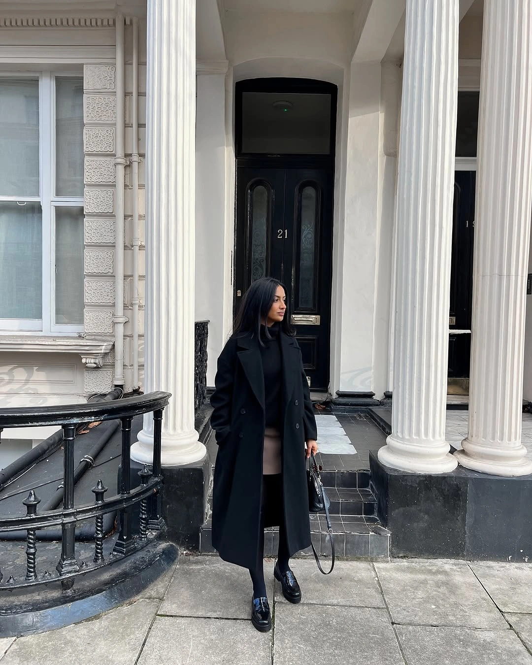 Long black coat with dark trousers, ankle boots, and pale shoulder bag