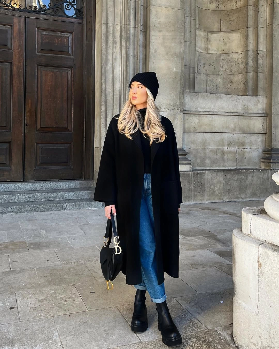 Long black coat with blue jeans, black boots, and knit hat