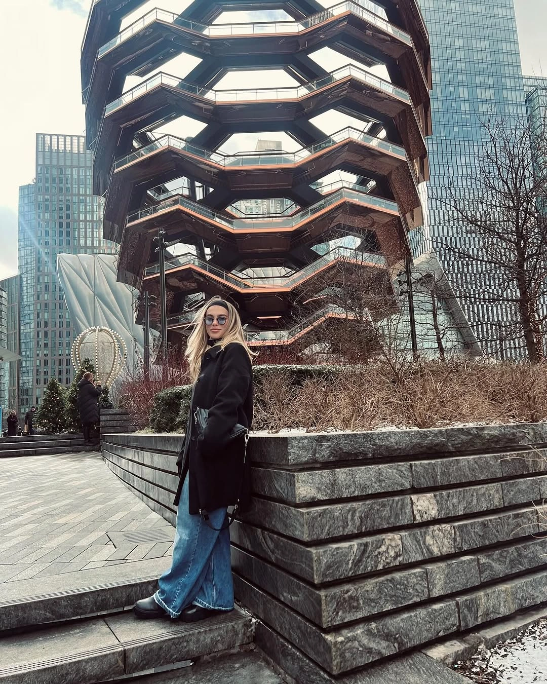 Woman in black coat and blue jeans near sculptural city building